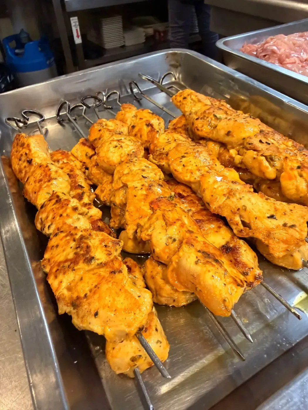 Brochetas de pollo sazonadas dispuestas en una bandeja metálica en una cocina comercial.