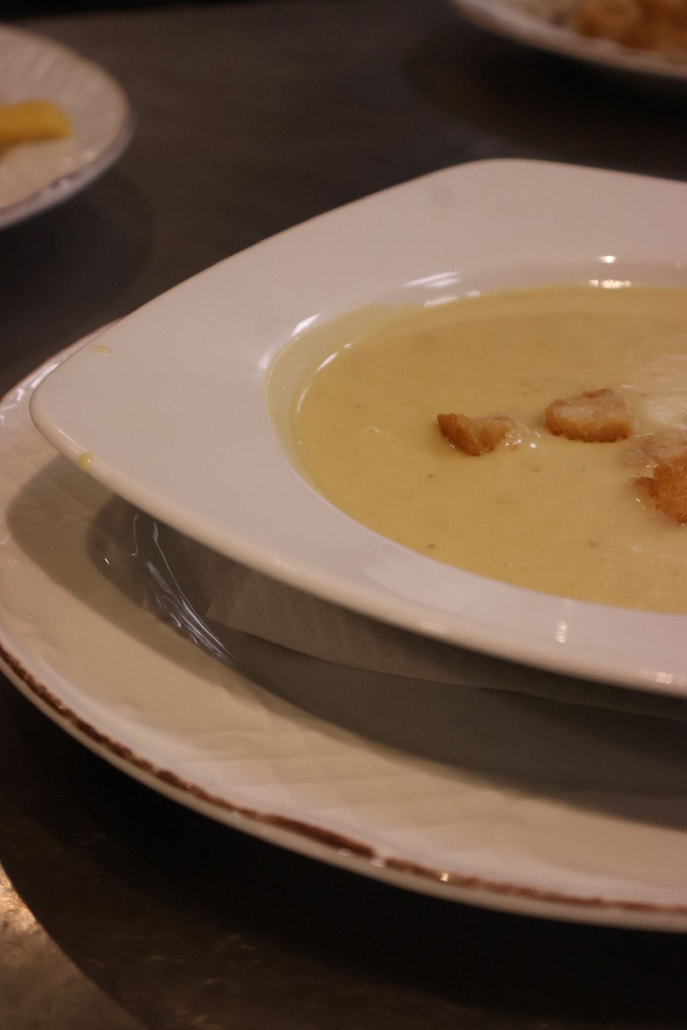 Un tazón blanco de sopa cremosa con picatostes sobre platos apilados en una mesa de comedor.