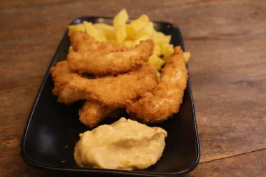 Un plato rectangular negro sobre una superficie de madera contiene tiras de pollo empanizadas.