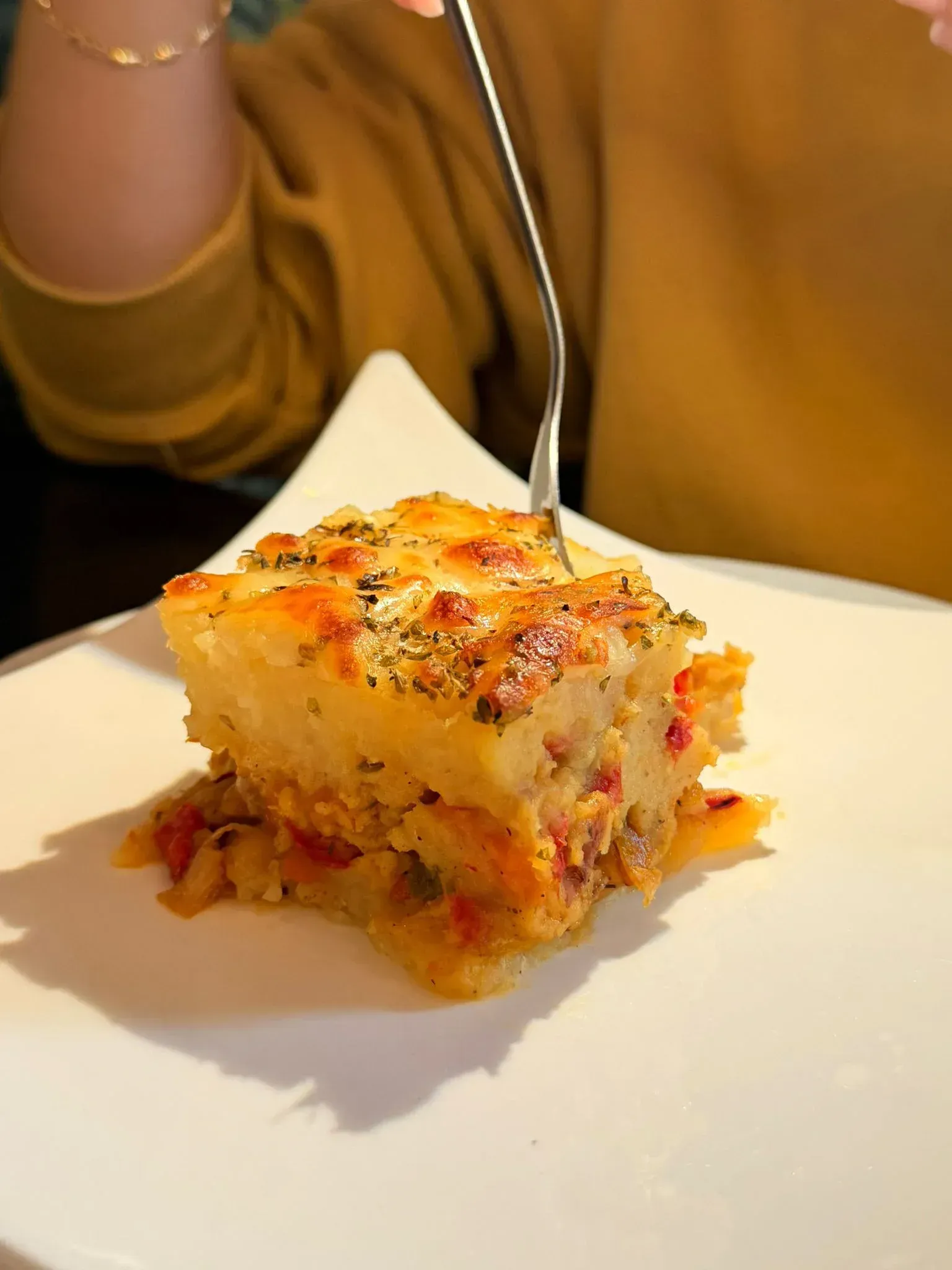 Una persona que lleva una camiseta amarilla usa un tenedor para comer un trozo cuadrado de un guiso salado horneado.