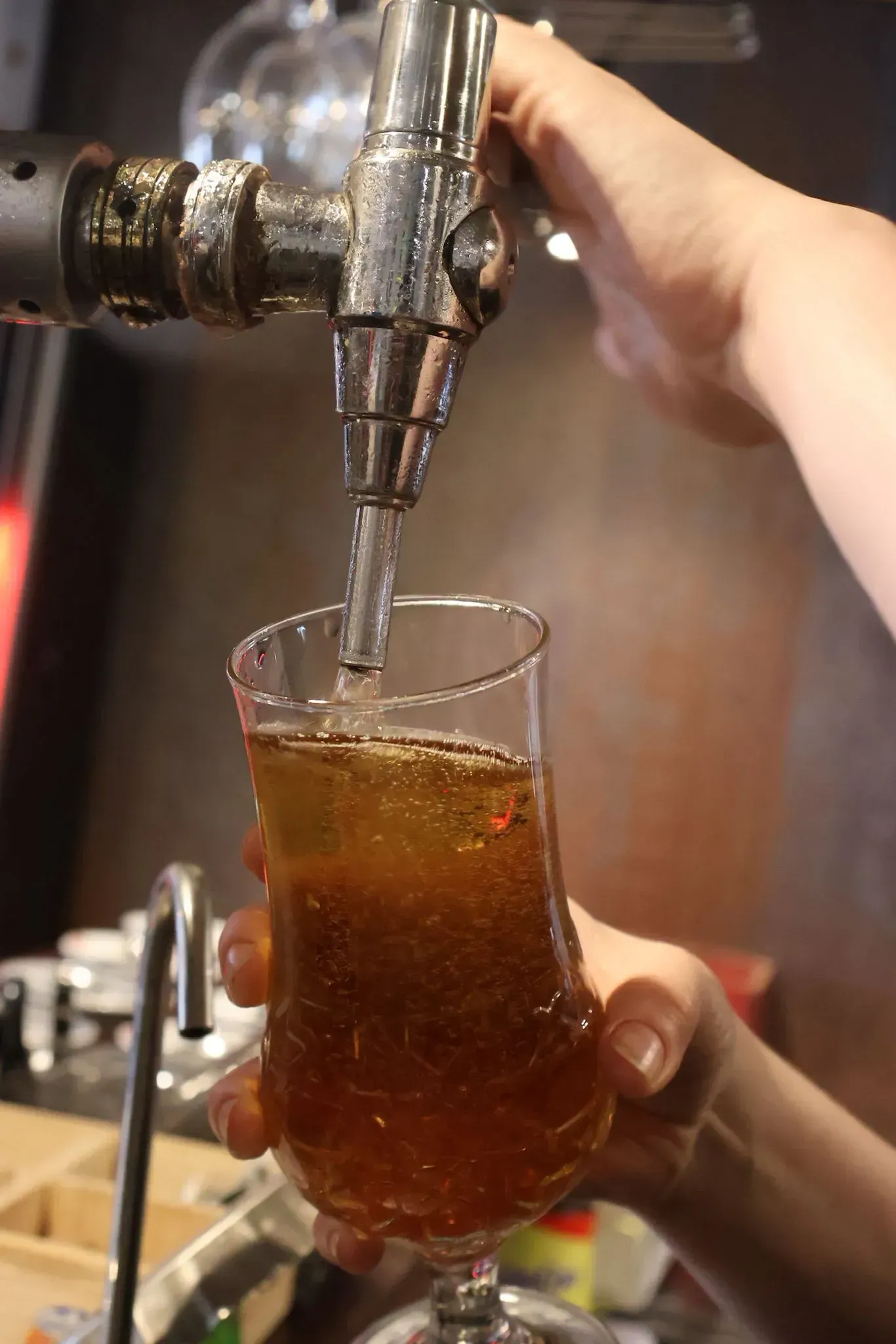 En un bar, una persona vierte con la mano una bebida dorada y carbonatada desde un grifo metálico a un vaso alto.