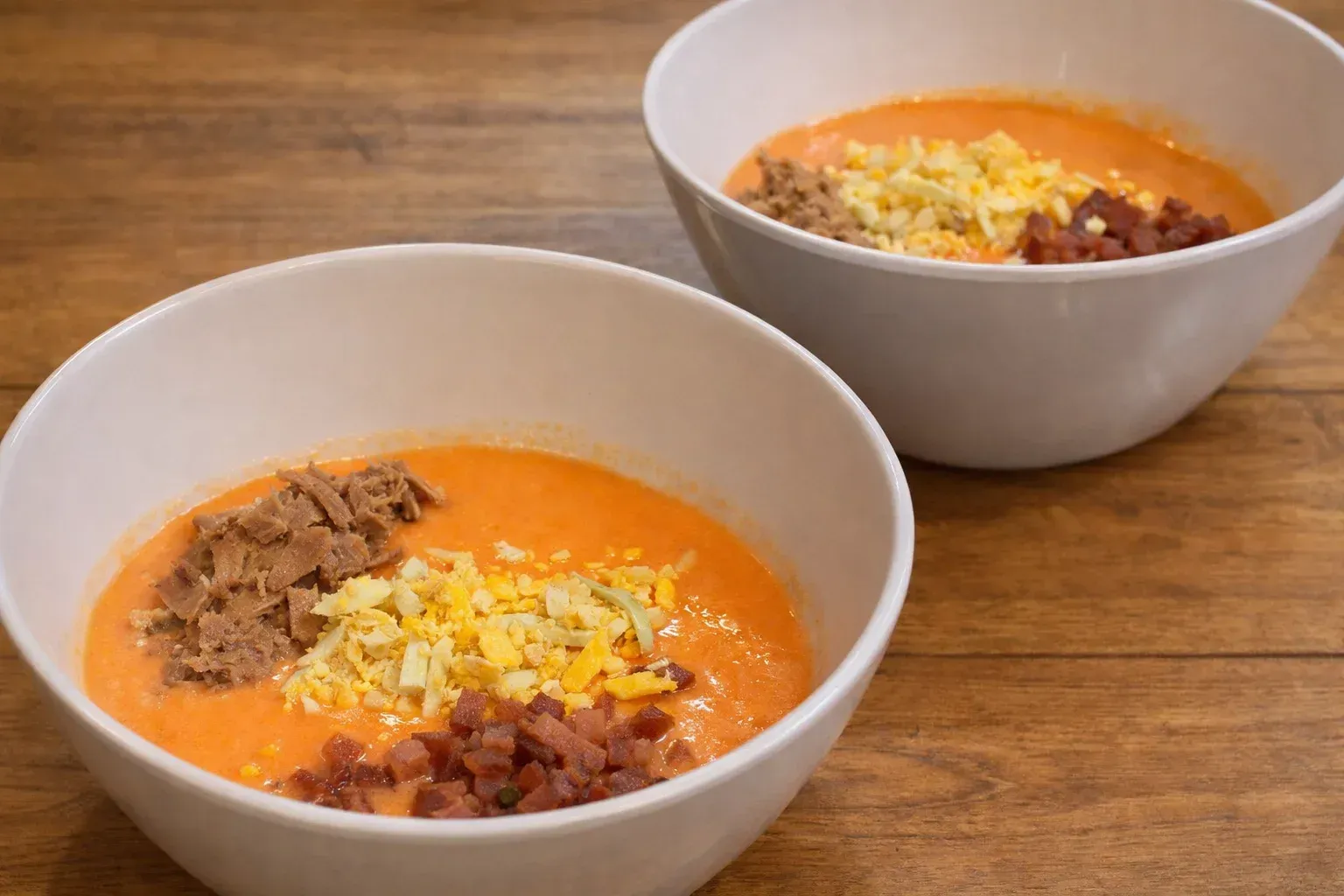 Two bowls of tomato soup topped with shredded cheese and crumbled meat on a wooden table