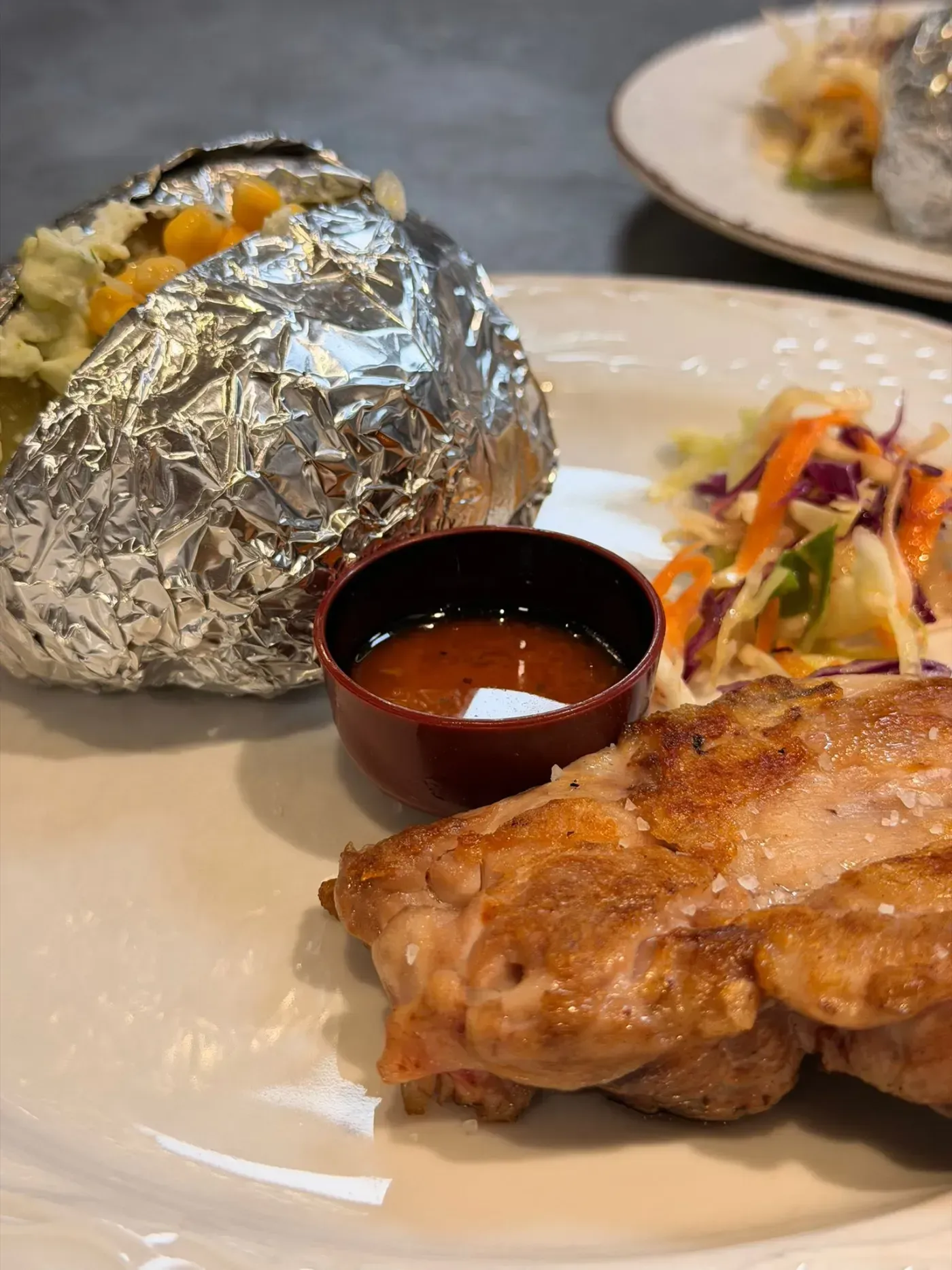 Un plato principal que incluye pollo a la parrilla, una patata asada envuelta en papel de aluminio y cubierta con maíz.