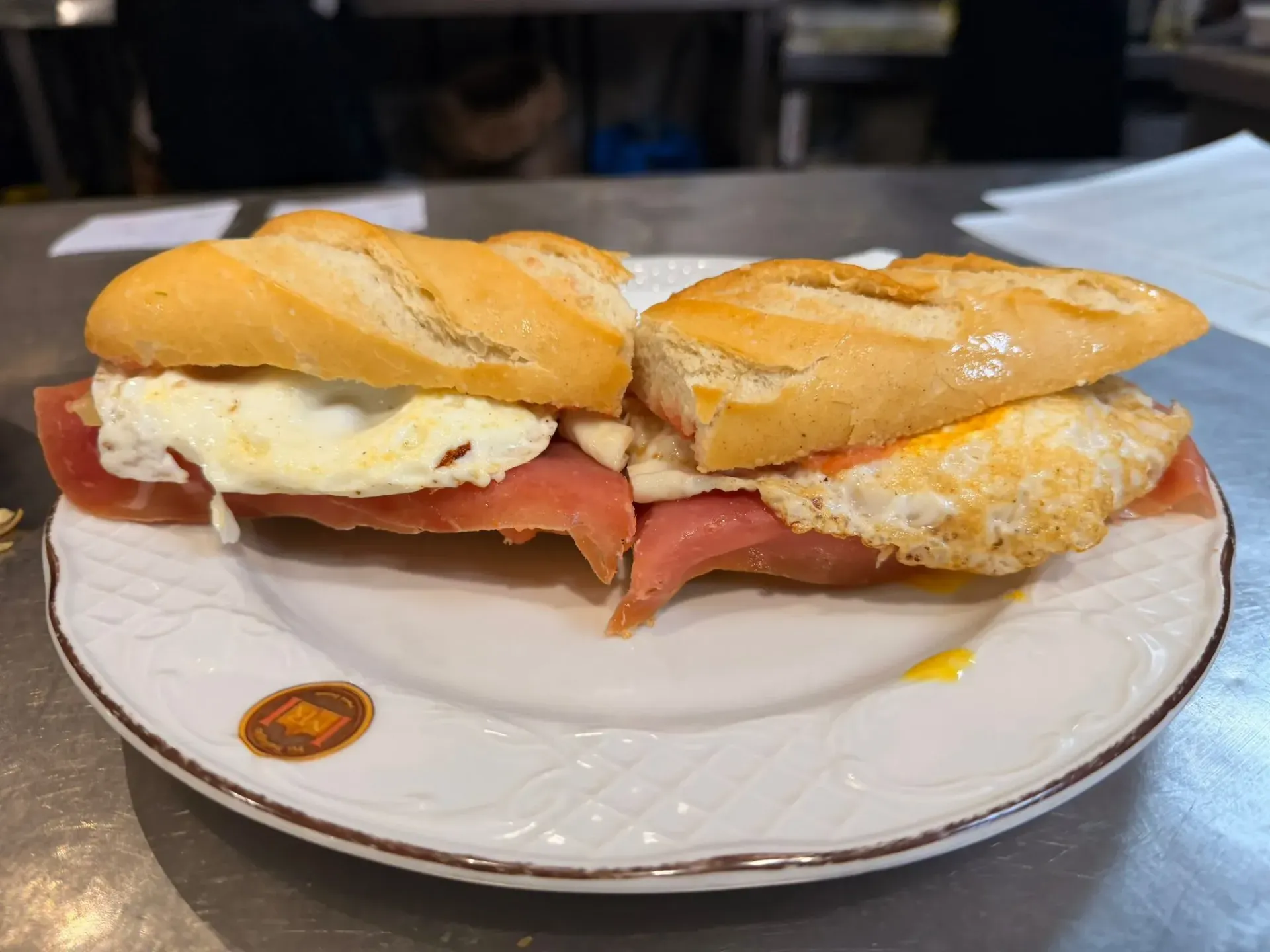 Un sándwich en un plato blanco, relleno de huevo, tomate y jamón, sobre la mesa de un café.