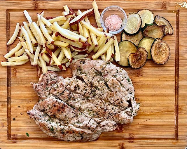 Una tabla de cortar de madera cubierta con carne, papas fritas y verduras.
