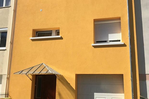 Façade d’un bâtiment en stuc orange avec deux fenêtres, un petit auvent et une porte d’entrée en plein soleil