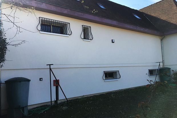 Côté d’une maison blanche avec des fenêtres à barreaux, une poubelle grise et une pelouse au crépuscule
