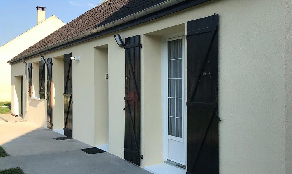 Extérieur d’une maison beige aux volets noirs et aux portes-fenêtres blanches, le long d’un mur latéral ensoleillé