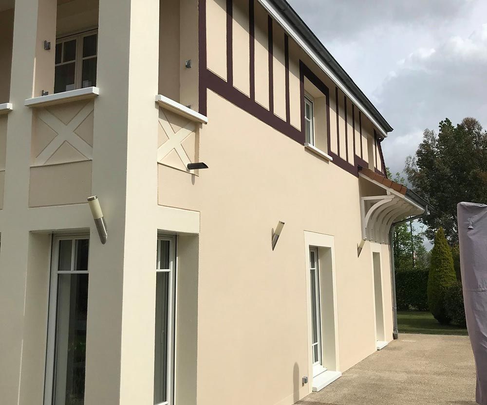 Extérieur d’une maison de deux étages couleur crème, avec des boiseries marron et une allée incurvée.
