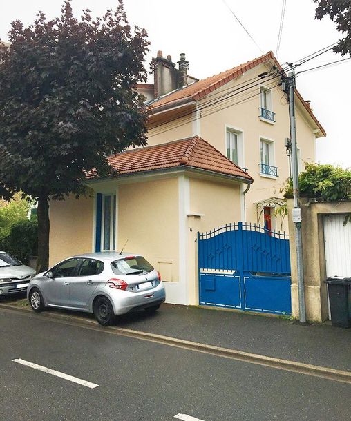 Maison couleur crème avec un toit rouge, un portail bleu et une voiture argentée garée dans une rue mouillée.