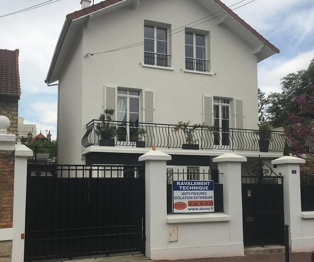 Maison blanche à deux étages avec portail et clôture noirs, balcon et panneau immobilier à l’entrée
