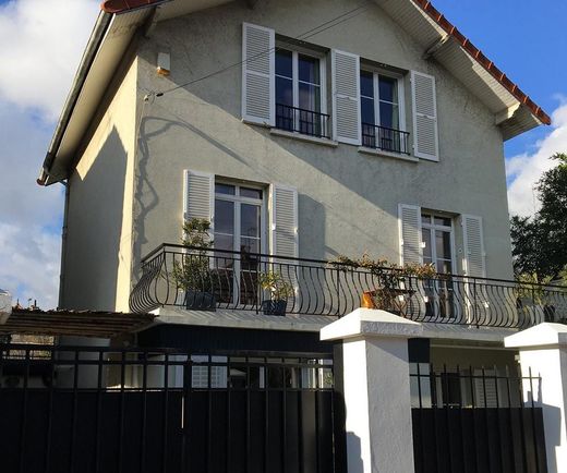 Maison crème à deux étages, avec des volets blancs, un balcon et un portail noir, sous un ciel partiellement nuageux