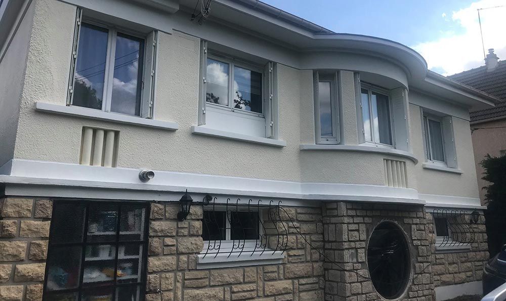 Extérieur d’une maison à deux étages avec un enduit crème, une façade inférieure en pierre et une fenêtre ronde près de l’entrée