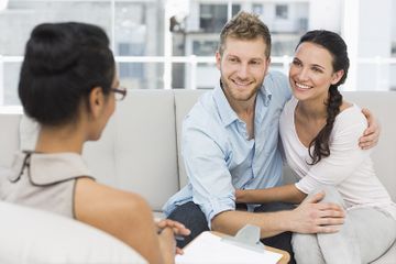 Una pareja sentada junta en una luminosa consulta de terapia mientras un terapeuta escucha.