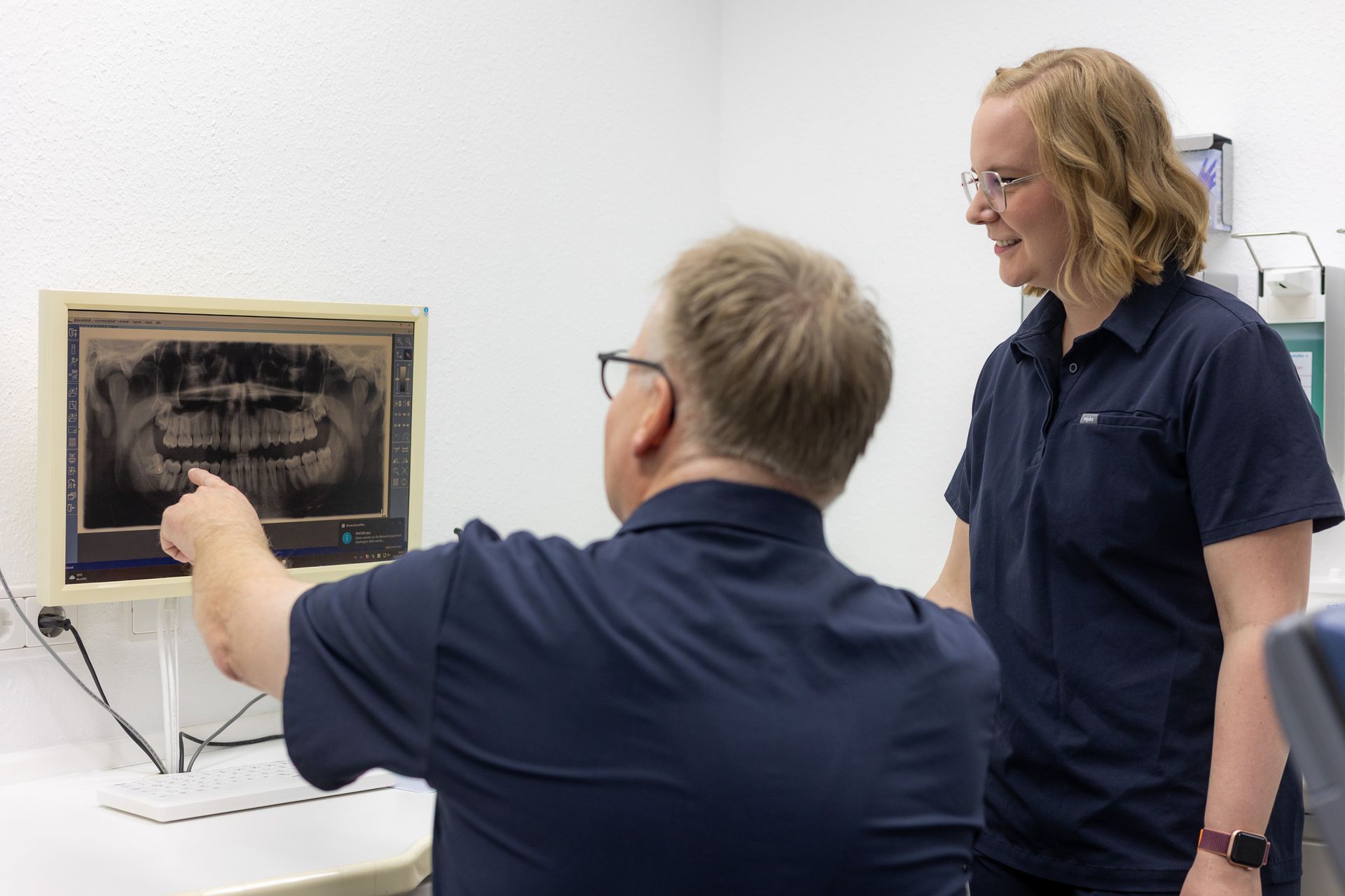 Der Zahnarzt zeigt auf ein Zahnröntgenbild auf einem Monitor; die Assistentin lächelt neben ihm in einer Zahnarztpraxis.