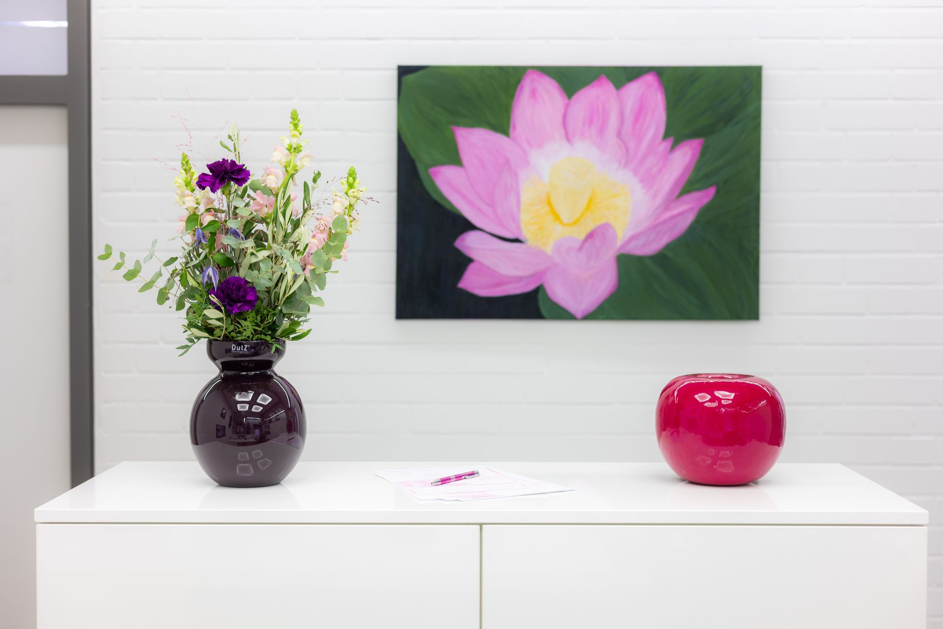 Weißer Schrank mit violetter Blumenvase, rosa Lotusgemälde und roter Kugel darauf.