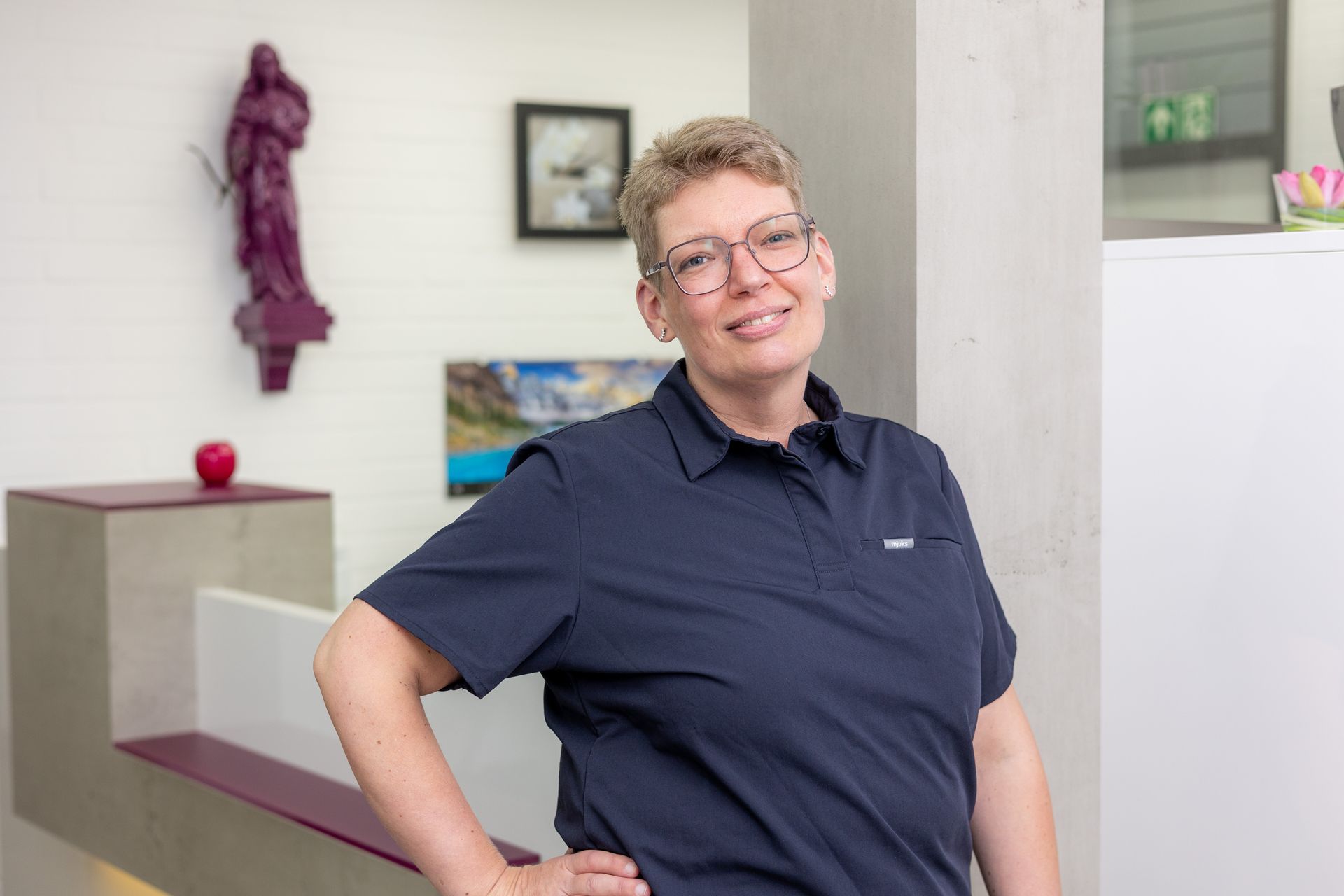 Frau mit Brille, dunkelblauem Poloshirt, lächelt. Lehnt sich an den Empfangstresen in einem Büro.