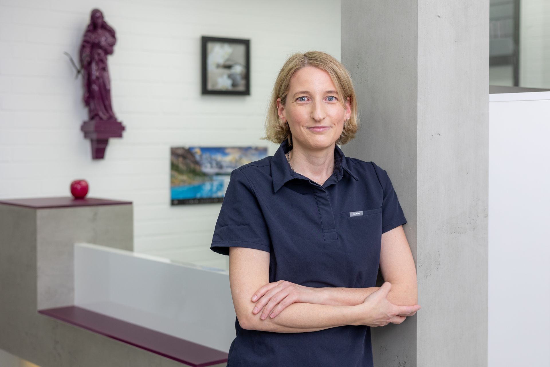 Frau in marineblauem Poloshirt, die Arme verschränkt, lehnt in einem Büro an einer weißen Wand.