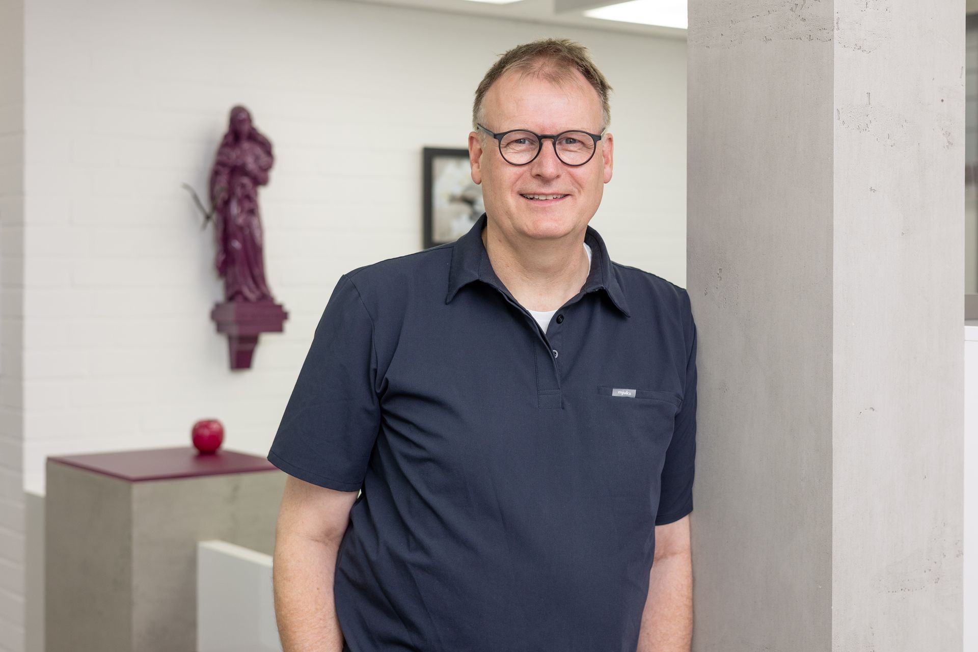 Lächelnder Mann mit Brille und dunkelblauem Hemd lehnt an einer weißen Wand. Im Hintergrund ein Kunstwerk.