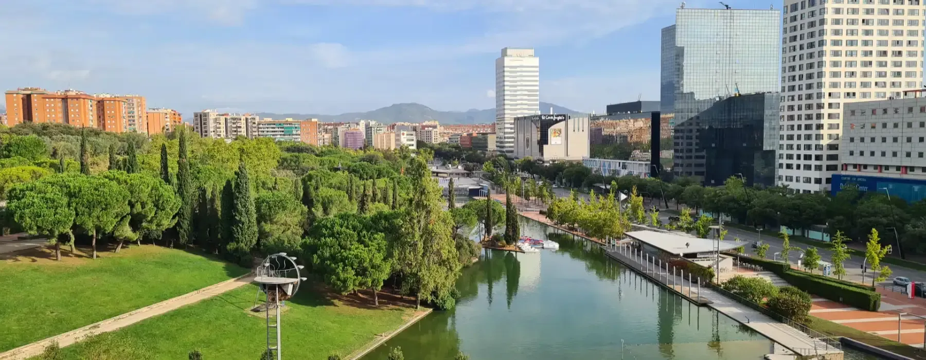 Un parque urbano con un río bordeado de árboles, un césped verde y edificios modernos bajo un cielo azul.