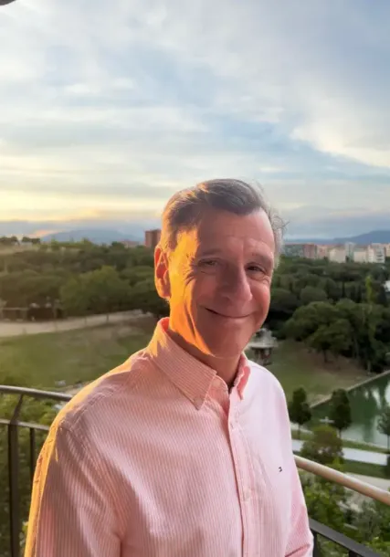 Hombre sonriente con camisa de rayas rosas en un balcón al atardecer con un paisaje pintoresco de fondo.