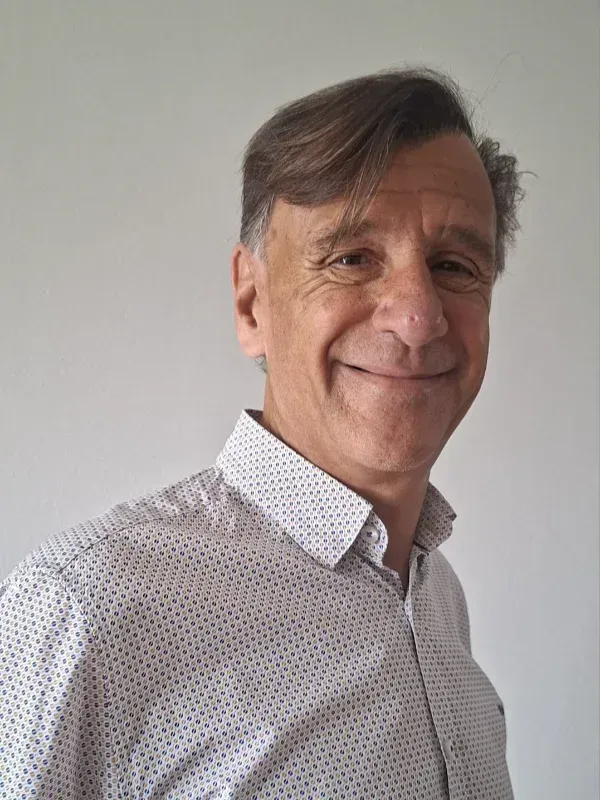 Hombre sonriendo con una camisa blanca estampada abotonada contra una pared lisa de color gris claro.