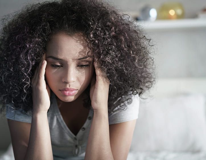 Mujer con cabello rizado, sujetándose las sienes, mirando hacia abajo con expresión angustiada.