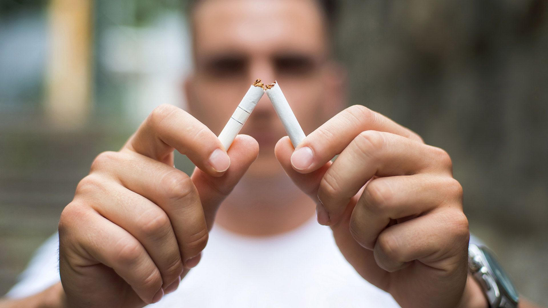 Hombre rompiendo un cigarrillo por la mitad; camiseta blanca, fondo borroso, foco en las manos y el cigarrillo.