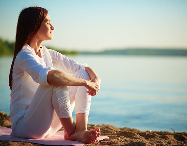 Mujer meditando en una playa, ojos cerrados, manos sobre las rodillas, expresión serena, día soleado.