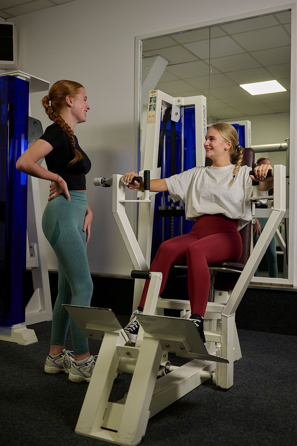 Eine Frau sitzt in einem Fitnessstudio auf einem Gerät, während eine andere Frau neben ihr steht.
