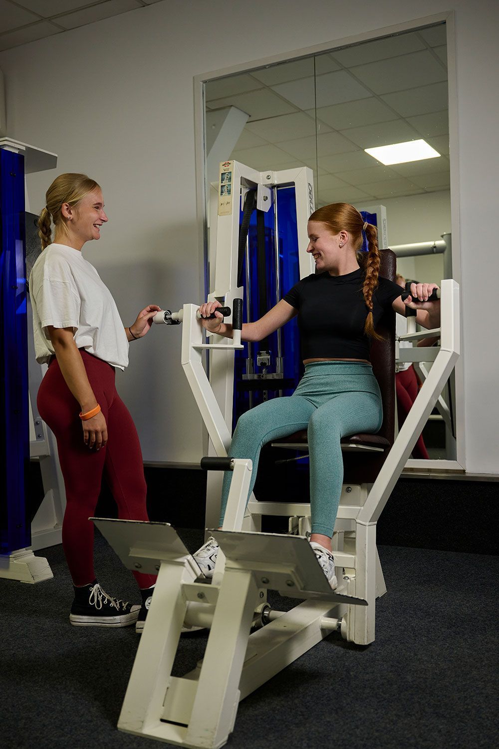 Eine Frau sitzt in einem Fitnessstudio auf einem Gerät und unterhält sich mit einer anderen Frau.