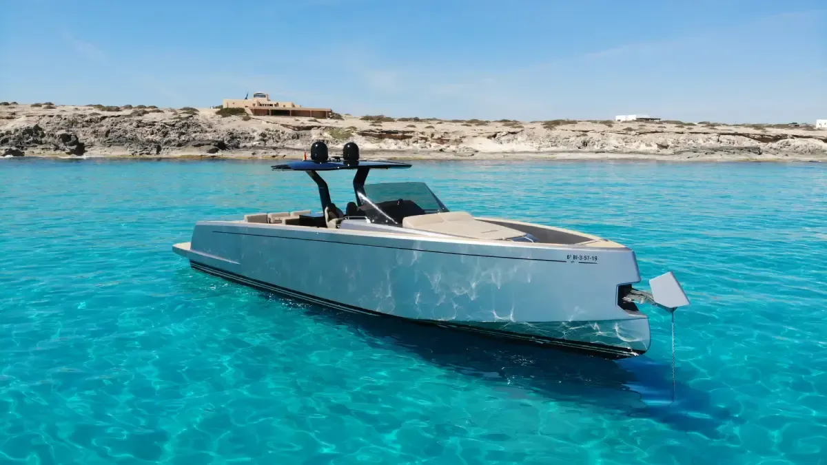 Yate a motor blanco en aguas de color turquesa brillante cerca de una costa rocosa bajo un cielo azul despejado.