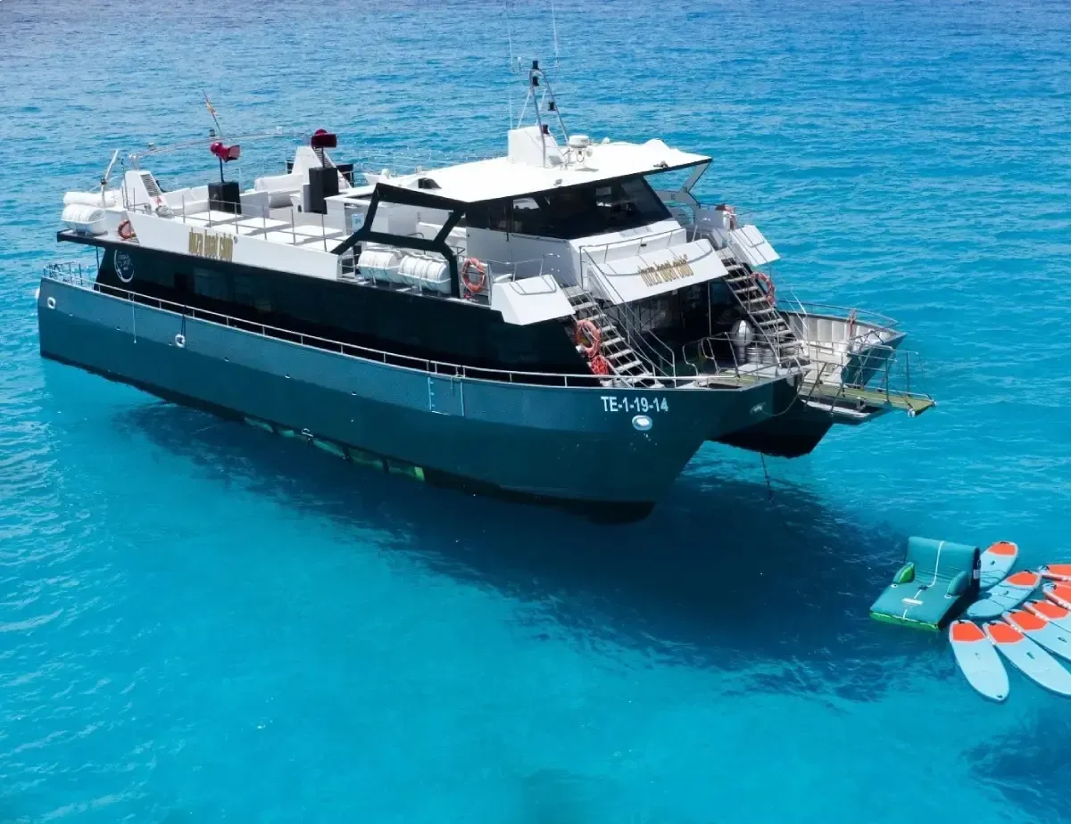 Un gran catamarán blanco y negro en aguas de un azul brillante, con una persona sobre una colchoneta inflable flotante cerca.