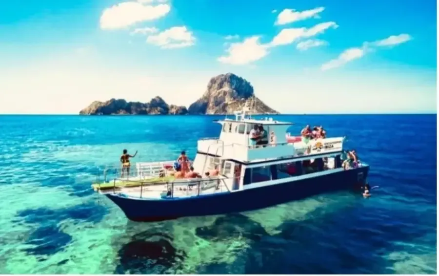 Barco turístico con gente en aguas turquesas cerca de una isla rocosa bajo un cielo azul brillante