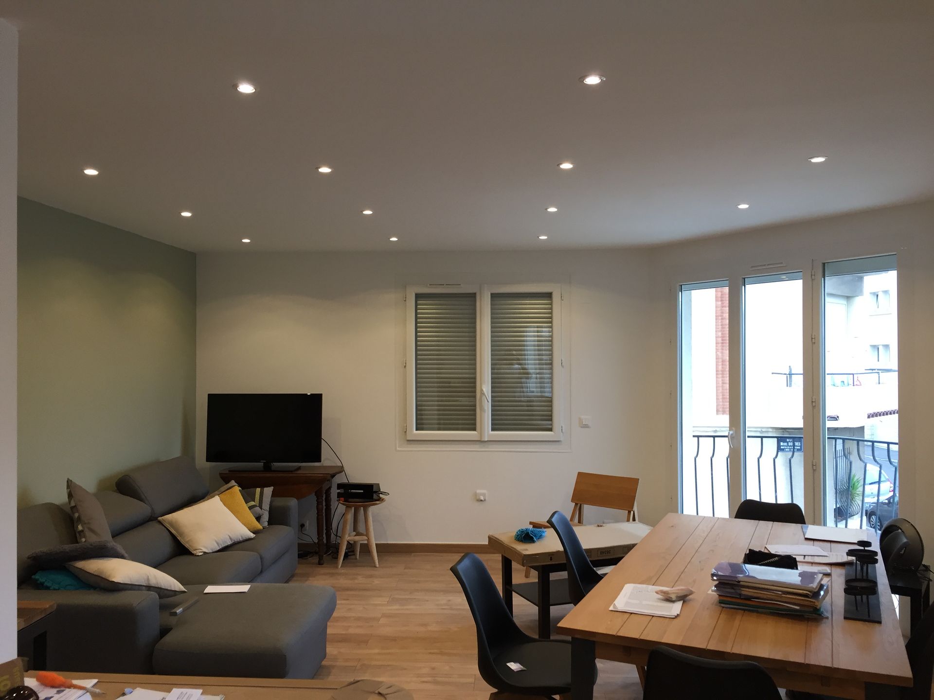 Salon avec canapé gris, télévision, table à manger et grandes fenêtres. Parquet clair, éclairage encastré.