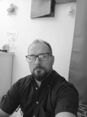 Homme portant des lunettes et une barbe, vêtu d'une chemise sombre, à l'intérieur.