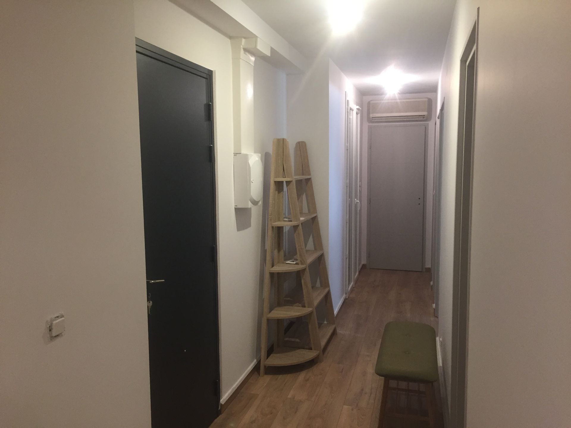 Couloir étroit au sol en bois. Une porte noire à gauche, une étagère en bois clair et un banc. Une autre porte se trouve plus loin.