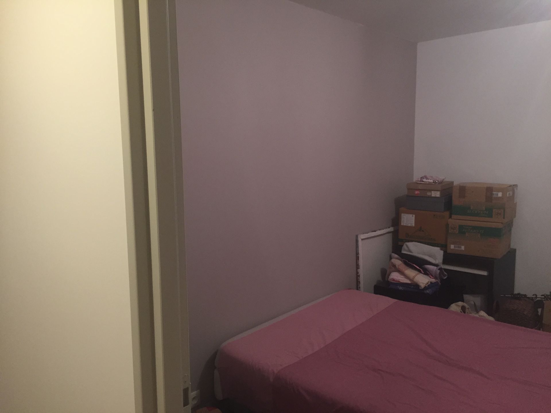 Chambre avec un lit, des boîtes et un mur peint en violet. Une porte se trouve à gauche.