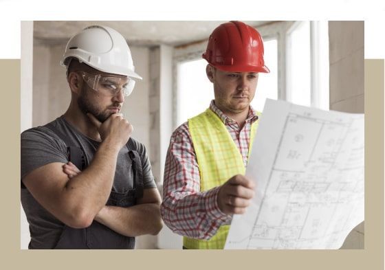 Deux ouvriers du bâtiment examinent un plan à l'intérieur d'un bâtiment, l'un portant un casque blanc et des lunettes de protection, l'autre un casque rouge et un gilet de sécurité.