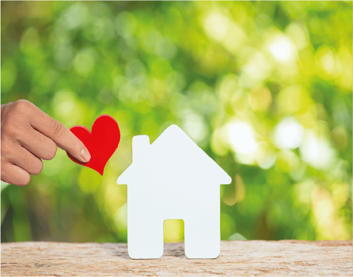 Une main dépose un cœur rouge à côté d'une silhouette de maison blanche sur une surface en bois à fond vert flou.