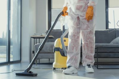 Eine Person im Schutzanzug saugt einen sonnendurchfluteten Raum in der Nähe eines grauen Sofas und eines gelben Staubsaugers.