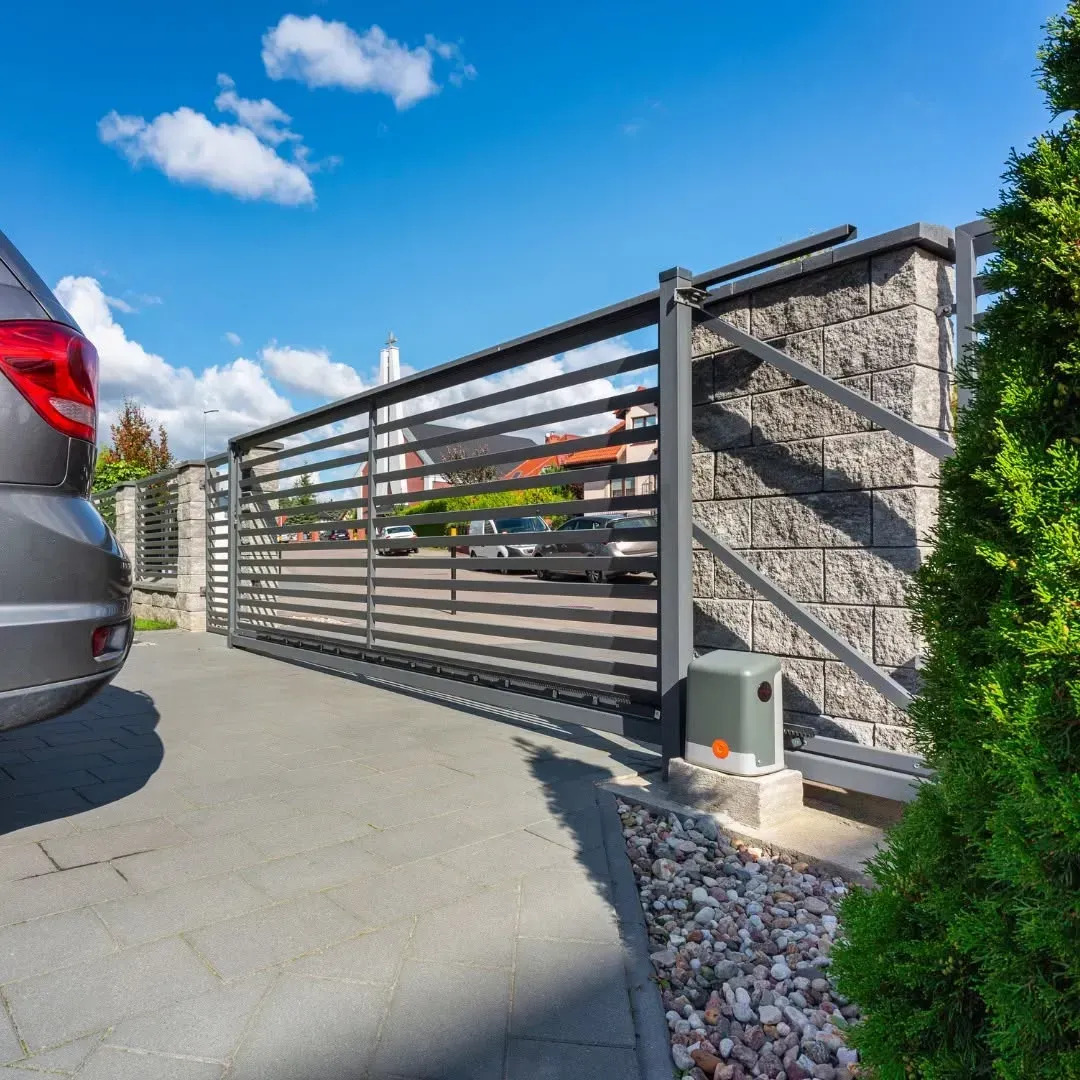 Coche gris cerca de una puerta metálica abierta, de listones horizontales, y de un muro de piedra en un día soleado.