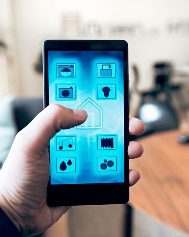 Mano sosteniendo un teléfono inteligente que muestra una aplicación de hogar inteligente con íconos de control; entorno interior borroso.
