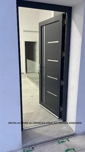 Puerta principal negra abierta con detalles plateados horizontales en un pasillo azul claro