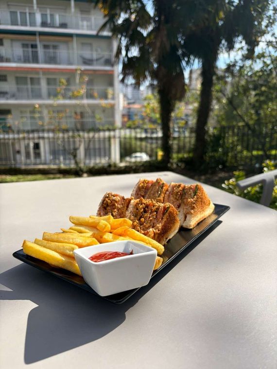 Un sándwich y papas fritas con un pequeño tazón de kétchup en una bandeja negra, dispuesta sobre una mesa al aire libre.