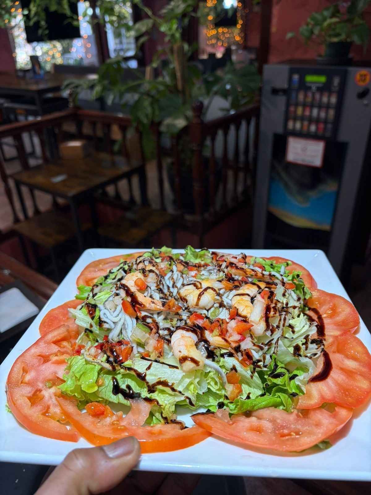 Un plato cuadrado blanco contiene una ensalada de camarones con lechuga, fideos de arroz y rodajas de tomate.