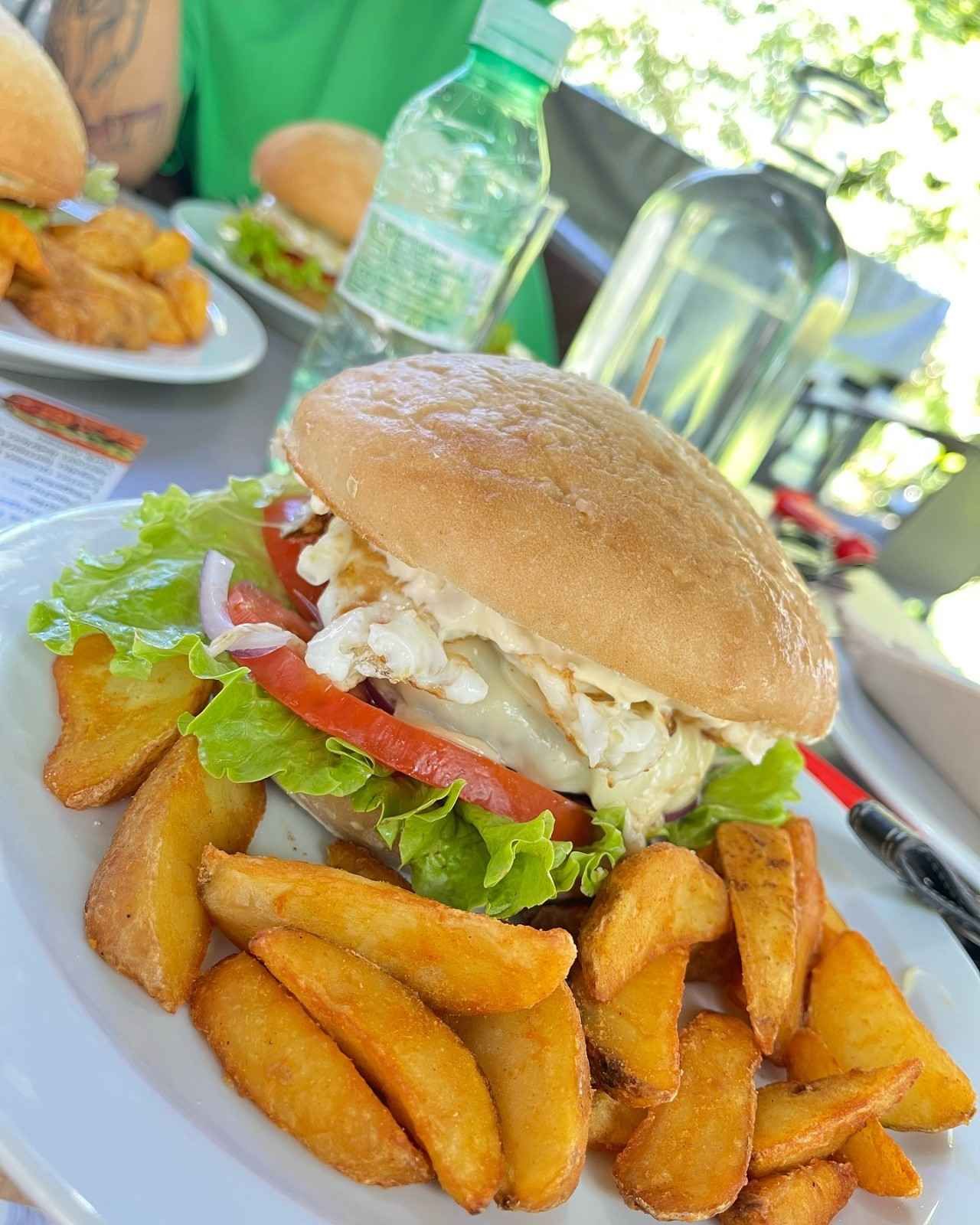 Una hamburguesa con lechuga, tomate y huevo frito en un plato blanco, servida con una generosa porción de patatas gajo.