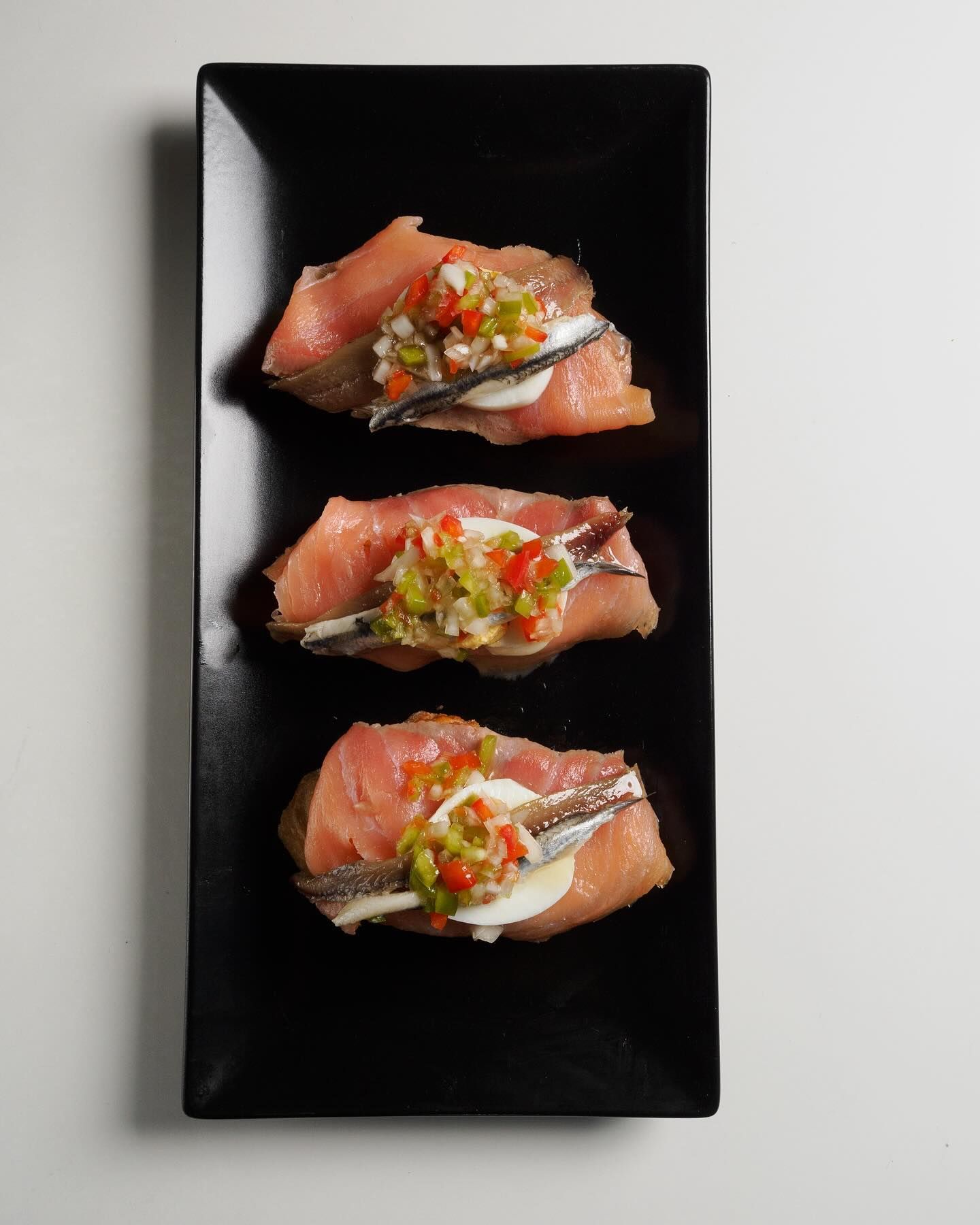 Tres canapés de salmón ahumado coronados con una anchoa, un huevo duro y una guarnición de pimientos picados.