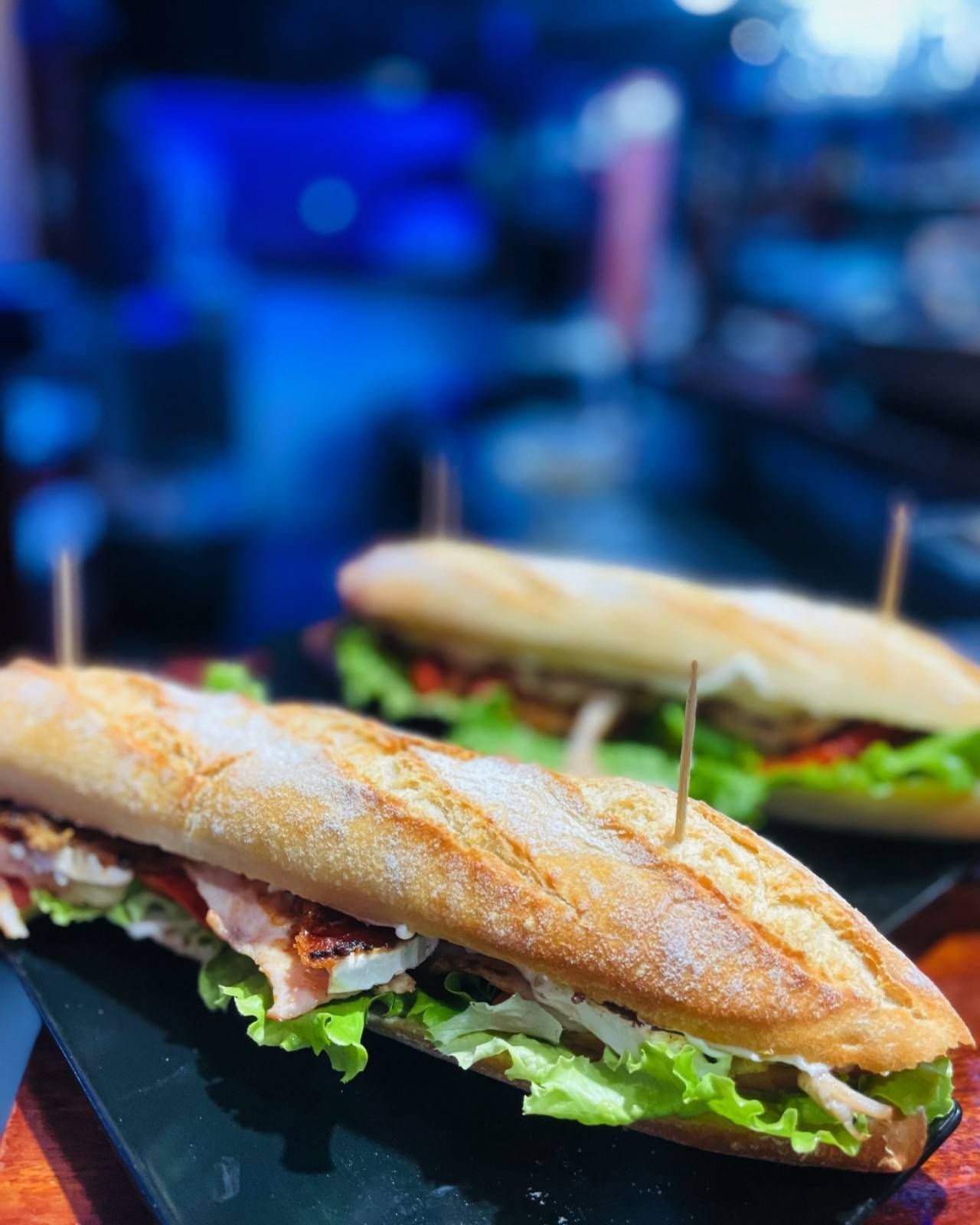 Dos bocadillos largos de baguette con lechuga y carne, servidos en una bandeja negra con palillos.