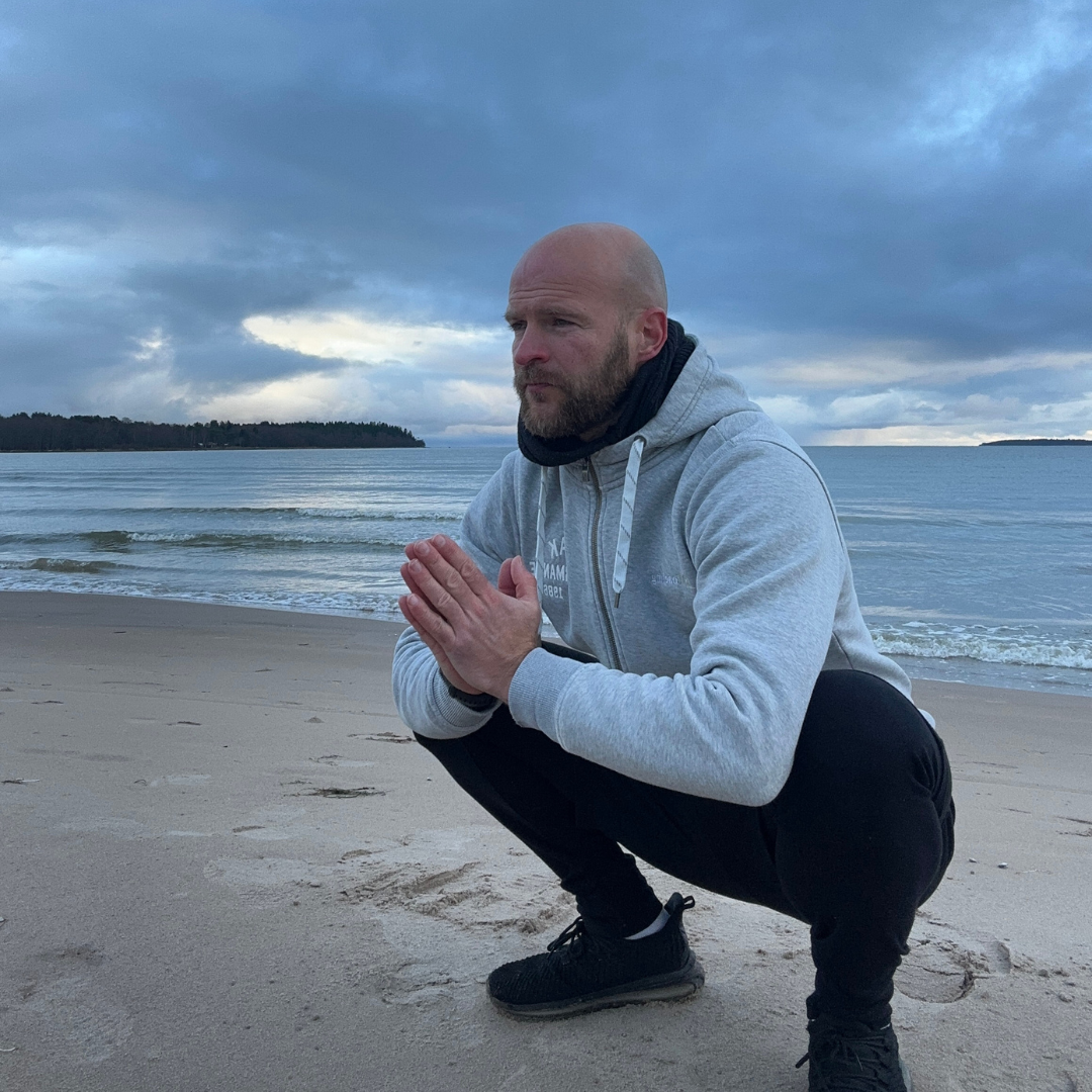 Harmaaseen huppariin pukeutunut mies kyykistyy rannalla kädet ristissä, tyyni vesi ja pilvinen taivas takanaan.